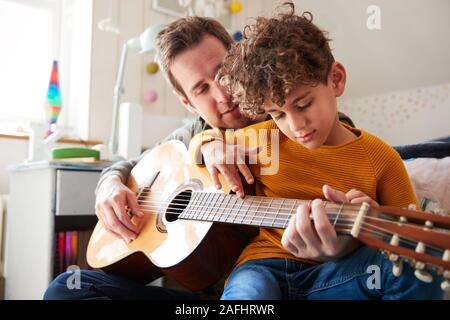 Single Father At Home With Son Teaching Him To Play Acoustic Guitar In Bedroom Stock Photo