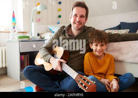 Portrait Of Single Father At Home With Son Playing Acoustic Guitar In Bedroom Stock Photo