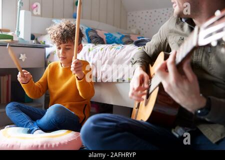 Single Father Playing Guitar With Son Who Drums On Cushion In Bedroom Stock Photo