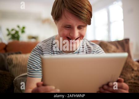 Young Downs Syndrome Man Sitting On Sofa Using Digital Tablet At Home Stock Photo