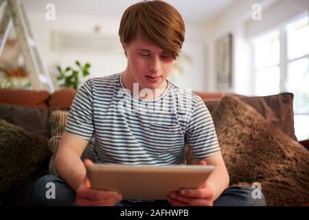 Young Downs Syndrome Man Sitting On Sofa Using Digital Tablet At Home Stock Photo