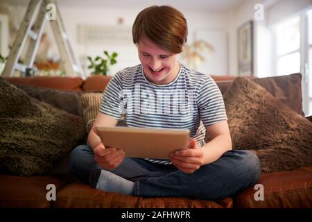 Young Downs Syndrome Man Sitting On Sofa Using Digital Tablet At Home Stock Photo