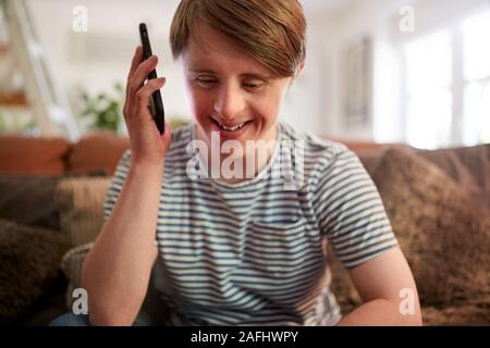 Young Downs Syndrome Man Sitting On Sofa Using Mobile Phone At Home Stock Photo