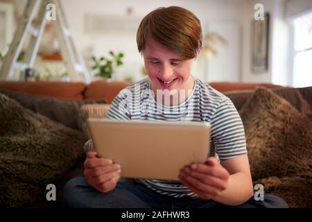Young Downs Syndrome Man Sitting On Sofa Using Digital Tablet At Home Stock Photo