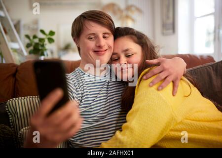 Loving Young Downs Syndrome Couple Sitting On Sofa Using Mobile Phone To Take Selfie At Home Stock Photo