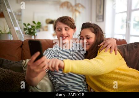 Young Downs Syndrome Couple Sitting On Sofa Using Mobile Phone To Take Selfie At Home Stock Photo