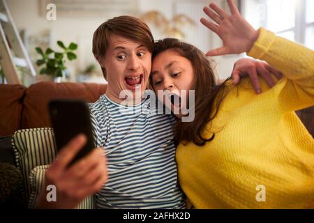 Young Downs Syndrome Couple Sitting On Sofa Using Mobile Phone To Take Selfie At Home Stock Photo
