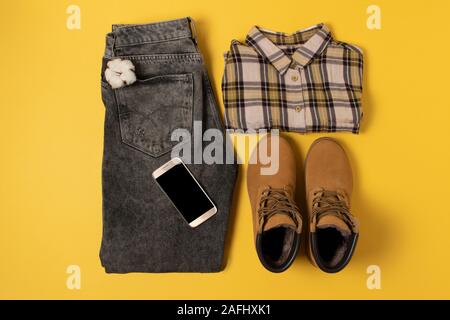 layout of autumn clothes and accessories in warm colors on a yellow background. Jeans, shirt, boots and phone flat lay nearby Stock Photo