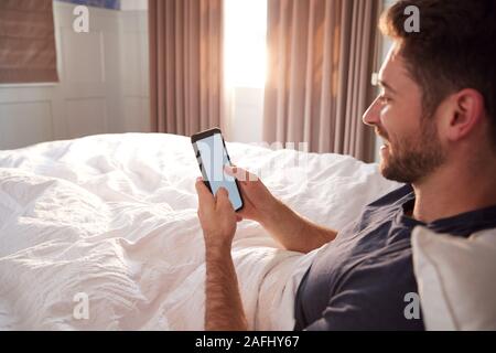 Man Sitting Up In Bed Looking At Mobile Phone After Having Woken Up Stock Photo