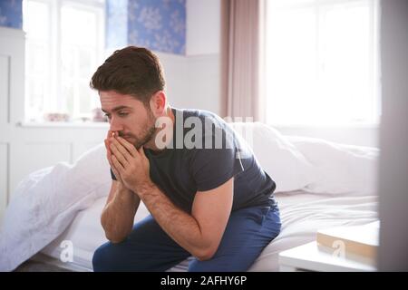 Man Wearing Pajamas Suffering With Depression Sitting On Bed At Home Stock Photo