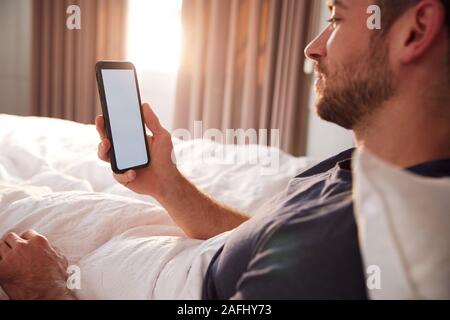 Man Sitting Up In Bed Looking At Mobile Phone After Having Woken Up Stock Photo