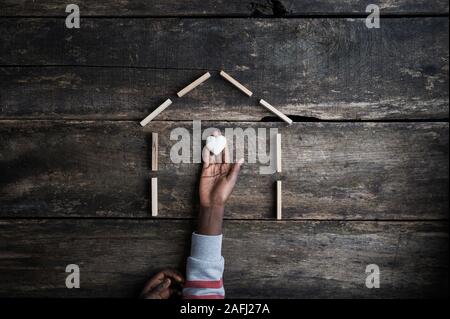 Hand of a colored girl holding heart shaped marble stone in a home made of wooden pegs and blocks. Over rustic wooden background. Stock Photo