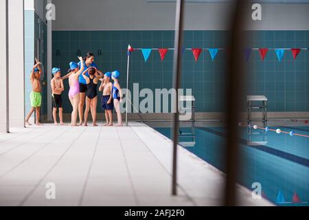 Female Coach Giving Children In Swimming Class Briefing As They Sit On Edge Of Indoor Pool