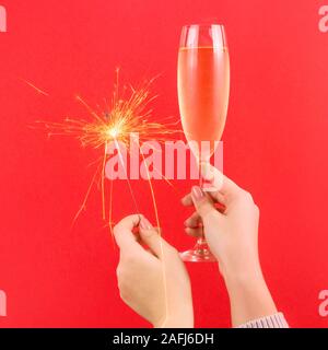 Female hand holds burning sparkler and a glass of champagne symbol of magic at Christmas on bright red background.
