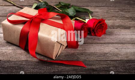 Closeup of Valentine gift wrapped in brown paper and red ribbon with two roses on rustic wooden surface Stock Photo