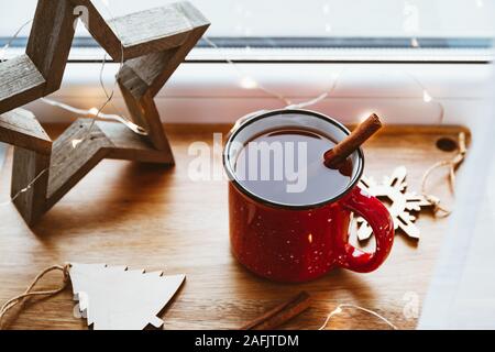 Black tea with cinnamon in a red mug among winter decor and lights. Cozy winter time still life. Stock Photo