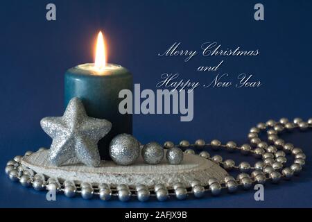 Silver and blue christmas composition with burning candle, glitter star, globules and bead chain on dark blue background - greetings text Stock Photo