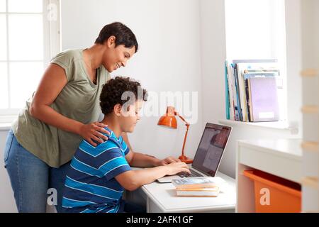 Mother Helping Son In Bedroom To Do Homework Using Laptop On Desk Stock Photo