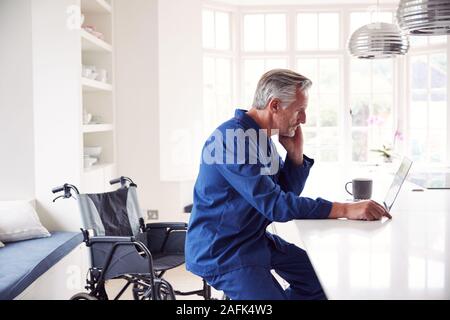 Mature Disabled Man In Wheelchair At Home Using Laptop On Kitchen Counter Stock Photo
