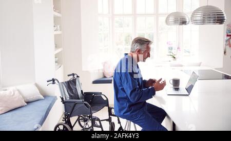 Mature Disabled Man In Wheelchair At Home Using Laptop On Kitchen Counter Stock Photo