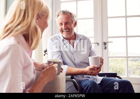 Mature Couple With Man In Wheelchair Sitting In Lounge At Home Talking Together Stock Photo