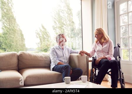 Mature Couple With Woman In Wheelchair Sitting In Lounge At Home Holding Hands Together Stock Photo