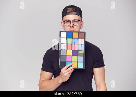 Portrait Of Male Photographer In Studio Holding Colour Checker Card Against White Background Stock Photo