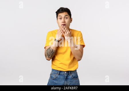 Scared, shocked young astonished asian guy in yellow t-shirt, gasping, cover opened mouth with palms and stare camera impressed, gossiping, realise Stock Photo