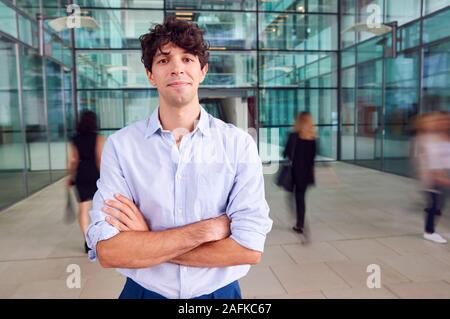 Portrait Of Young Businessman With Crossed Arms Standing In Lobby Of Busy Modern Office Stock Photo