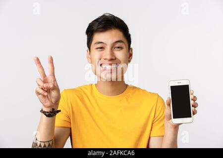 Close-up shot attractive enthusiastic, joyful asian man in yellow t-shirt, showing peace sign and smartphone screen, promote application, messanger or Stock Photo