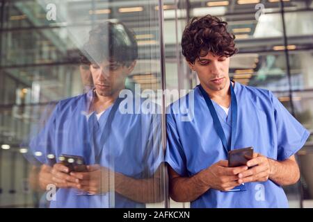 Young Male Doctor Wearing Scrubs Using Mobile Phone Leaning Against Wall In Modern Hospital Building Stock Photo