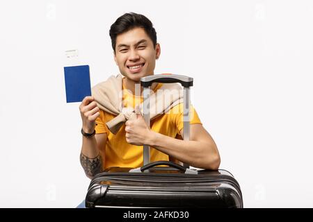 Cheeky and happy asian, chinese man in yellow t-shirt, sitting black suitcase, luggage and thumb-up pleased, holding passport with plane ticket Stock Photo