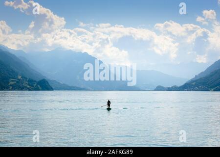 Silhouette of a man standing on a paddleboard on Lake Brienz in Switzerland. Hills in the background. Active lifestyle, sport activities. Peaceful background, motivational concept. Stock Photo
