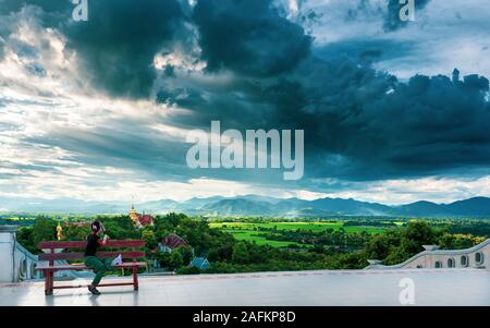 Traveler woman enjoy scenery nature landscape beautiful Thailand temple on background rice field and mountain in fog on horizon at sunset in Asia. Stock Photo