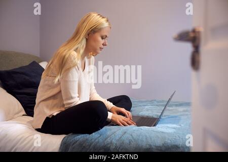 Female College Student Sits On Bed In Rented House Using Laptop Stock Photo