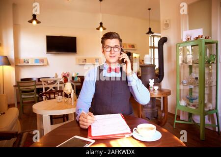 Shot of young cafe owner man wearing bow tie and apron while sitting at table making a call. Stock Photo