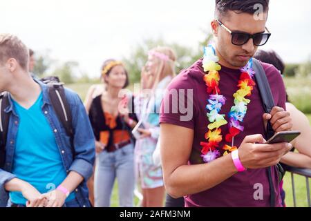 Young Man Looking At Mobile Phone As He Waits Behind Barrier At Entrance To Music Festival Site Stock Photo