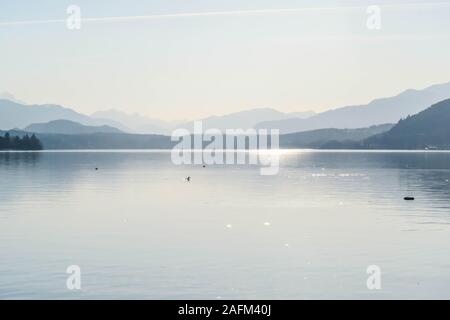 A view on a lake and Alps in the back. The calm surface of the lake is reflecting the mountains, sunbeams and clouds. Clear and sunny day. Calm and re Stock Photo