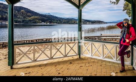 Girl wearing pink jacket leans on a balustrade of a pavilion. She is looking straight at the beautiful Wörter Lake, located at the bottom of the Alps. Stock Photo
