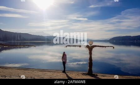 A girl in a winter jacket walks along the lakeside. There is a scarecrow next to the shore. The surface of the lake is still and calm. There are tall Stock Photo