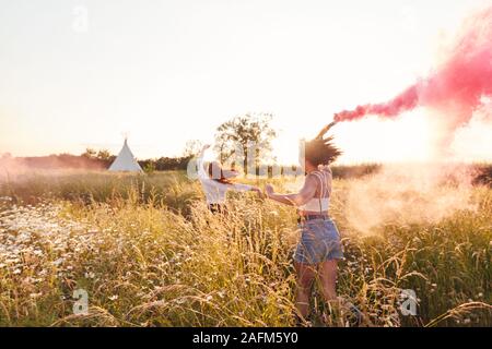 Two Female Friends Camping At Music Festival Running Through Field With Smoke Flares Stock Photo