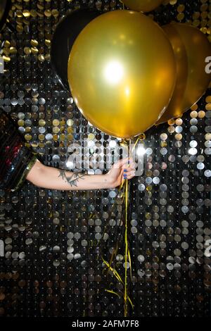 Hand of young woman with tattoo holding golden balloon against glittering wall Stock Photo