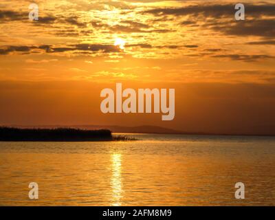 Sunset over the lake. Sun goes down behind the clouds. Sunbeams reflecting in the calm surface of the lake. A small island in the middle of the lake. Stock Photo