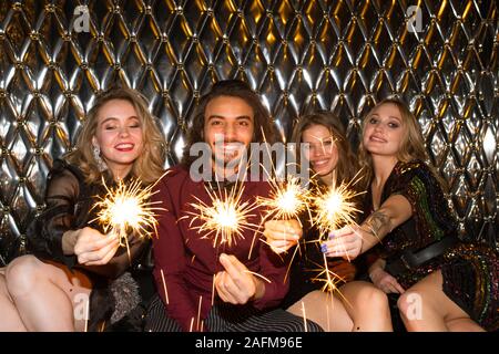Happy girls and guy with sparkling bengal lights sitting by wall in night club Stock Photo