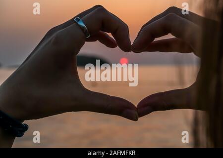 A heart formed from hands, and framing the setting sun in it. The sun sets over the horizon. The sun beams reflecting in the calm sea waters. The sky Stock Photo