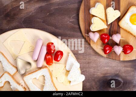 Cooking, sandwiches, heart-shaped snacks, top view. Heart-shaped sausages and sandwiches with cherry tomatoes with croutons on a wooden table Stock Photo