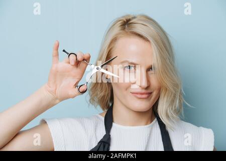 Stylish blonde female hairdresser holding open scissors next to her face Stock Photo