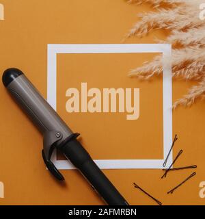 Composition of hairdresser tools: curling iron and bobby pins next to fluffy ears of cereal bouquet. Over orange background with white square on it. T Stock Photo