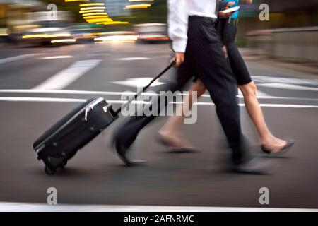 Two business people walk across a street, one carrying luggage. Stock Photo