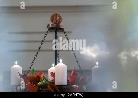 An Advent wreath hung with chains on a metal rod. It is made of branches, red leaves and flowers. Four white candles burn. There is smoke above them. Stock Photo